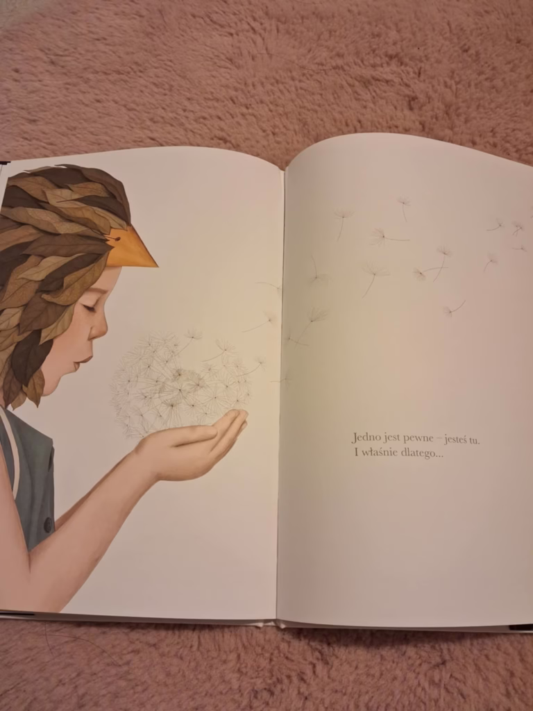 Wiara w siebie – dziecko w przebraniu z liści dmucha na dmuchawiec, którego nasiona unoszą się w powietrzu. Ilustracja z książki „Może” Kobi Yamady, symbolizująca marzenia, delikatność i poczucie własnej wartości.