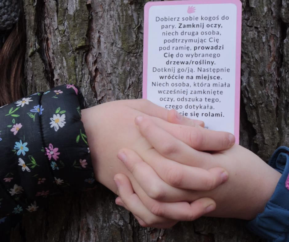 Dwoje dzieci trzymających się za ręce przy drzewie, wykonujących ćwiczenie z karty Dzieci w Naturze polegające na rozpoznawaniu rośliny dotykiem z zamkniętymi oczami.